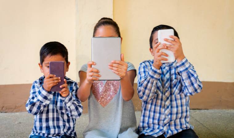 3 children sitting on a couch and all looking at phones and tablets, to illustrate that even young children are prey to sexual grooming